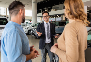 Young car salesman showing to mid-age couple new automobile at dealership salon.