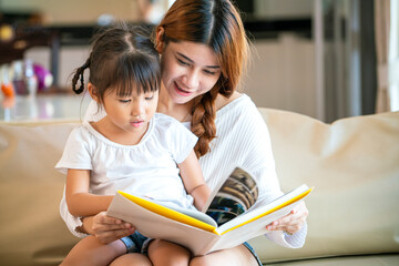 Asian pretty mother and her happy daughter reading story book together on cozy sofa in living room...