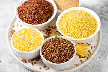 White and red rice, buckwheat, amaranth seeds, corn groats, quinoa and millet in white bowls on the light gray kitchen table. Gluten-free cereals	