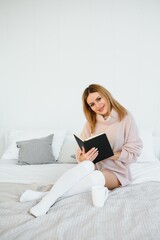 A woman reading a book and smiling as she sits in bed. The alarm clock on the desk beside her.