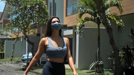 European brunette sports girl walks along an empty city street with green trees on the background alone in a gray protective mask