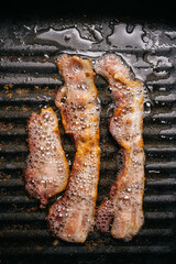 Top view of frying sizzling bacon strips in a pan with hot greasing bacon oil for making crispy bacon. Keto snack meal.