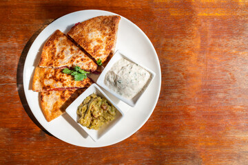 Top view of Vegan Quesadilla: grilled whole-wheat tortilla filled with sweet potato and red bean, vegan quesso cheese served with guacamole salsa and Tzaziki sauce.