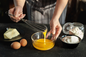 The process of preparing the dough. The woman whips the egg mixture with a whisk against a dark background. Ingredients for cookies.