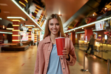 Portrait of lovely young woman smiling at camera, holding a soda, posing in front of a concession stand in a movie theater