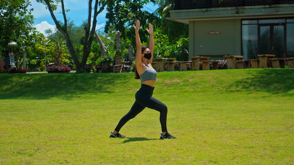 European brunette girl practices alone yoga in nature standing on green grass wearing a black protective mask