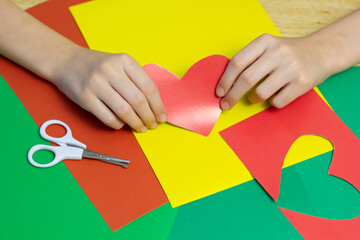 
The child cuts red hearts out of paper with scissors and makes a gift. Close-up. Colored background. Place for an inscription.