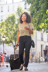 Full body smiling young woman traveling with bag and mobile phone in city