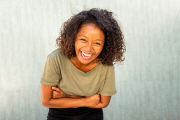 laughing young African American woman with curly hair and arms crossed by green background