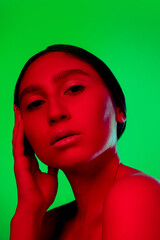 Serious. Beautiful east woman close up portrait isolated on green background in red neon light. Details, skin care and make up. Concept of human emotions, facial expression, sales, ad, fashion and