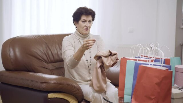 Shocked Caucasian woman looking at expensive dress price sitting on couch at home. Portrait of middle-aged shopaholic regrets on buying. Shopaholism and fashion concept.