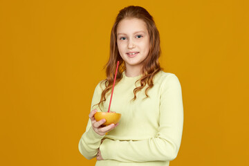 Fototapeta na wymiar cute little child girl holding fresh juicy grapefruit and drinks juice isolated over yellow background