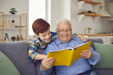 Older family members and children having fun together: Happy grandfather and grandson reading book of good fairy tales on cozy couch at home. Senior man and little kid enjoying free time on weekend