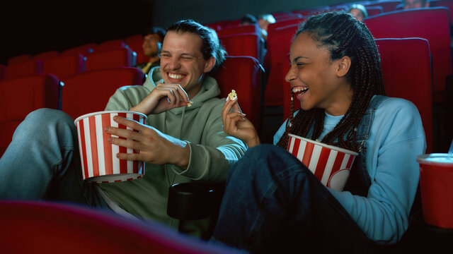 Cheerful best friends laughing, having popcorn while watching movie together, sitting in cinema auditorium