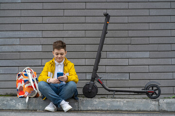 Teenager with mobile phone sitting next to his electric scooter and backpack. Modern young boy...