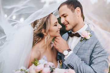 A young groom in a gray suit and a cute, smiling blonde bride with a bouquet are hugging tenderly...