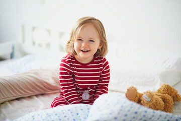 Happy toddler girl in striped red and white pajamas sitting on bed right after awaking