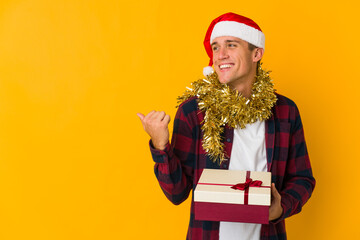 Young caucasian man with christmas hat holding a present isolated on yellow background