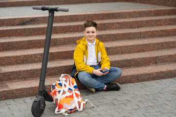 Modern teenager sits on the stairs with mobile phone in hands near his electric scooter