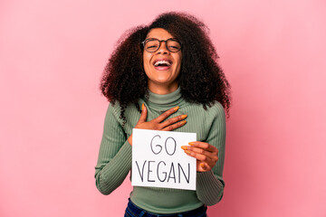 Young african american curly woman holding a go vegan placard laughs out loudly keeping hand on chest.