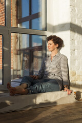 An active, beautiful middle-aged woman with a beautiful smile. Sitting on the windowsill near the window. Reflection in the window.