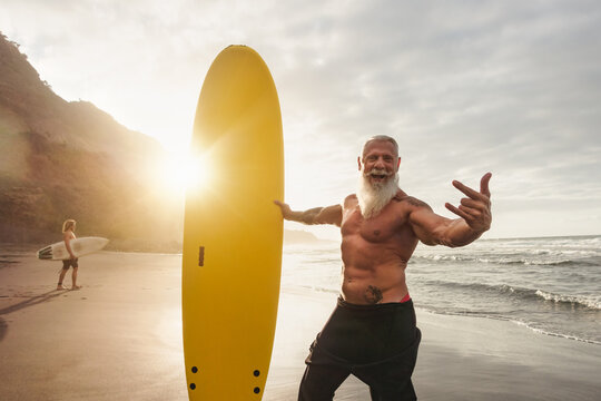 Happy fit senior having fun surfing at sunset time - Sporty bearded man training with surfboard on the beach - Elderly healthy people lifestyle and extreme sport concept