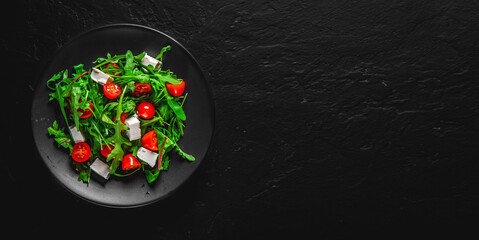 Fresh salad with tomatoes, arugula and cheese cubes on black plate on Dark grey black slate background