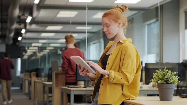 Medium slow-motion shot of young red-haired female office worker in bright yellow shirt working on tablet standing in big spacious office, while coworkers communicating in background