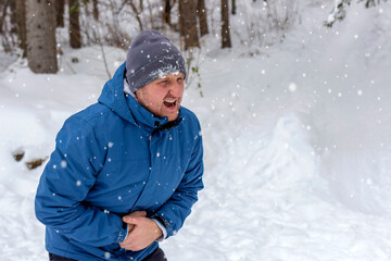 Fototapeta na wymiar Young handsome caucasian man wearing cap and jacket on snowy weather with hand on stomach causing pain in stomach, feeling unwell. Photo of young man with stomach pain. Frustrated man with stomachache