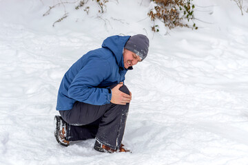 Fototapeta na wymiar Photo of injured young man bend down on snow and holding his knee in pain, outdoor. Young man holding his knee in pain on a snowy cold winter day. Man having a knee injury on winter road. Copy space.