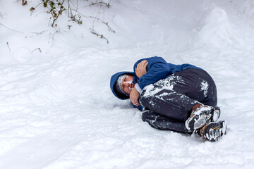 Photo of a frozen young man in blue jacket lying on the winter forest, covered snow. Man mountaineer lying ill with hypothermia on snow hill in winter. Bearded sick man frozen lies covered snowflakes.