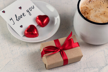 A gift with a red ribbon, a mug of coffee, heart-shaped chocolates and a note with the word I love you. Valentine's day breakfast.