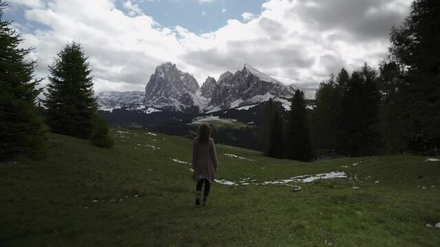 girl in brown dress slowly walks towards the Dolomites on a grass field