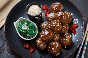 Black plate with roasted pork meatballs in teriyaki sauce and seaweed salad, close-up, studio shot