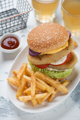 White plate with double fishburger, french fries and beer in the background, vertical shot on a white concrete surface