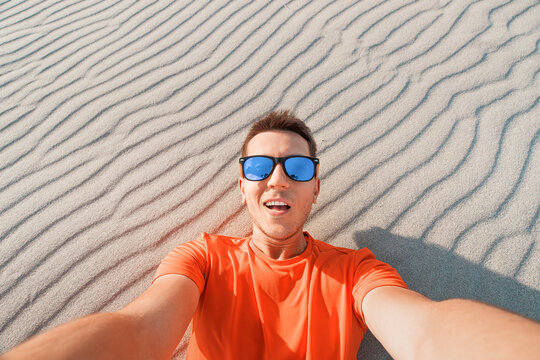 Happy man tramp in sunglasses takes a selfie photo in the desert against the background of a dune of white sand
