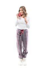 Joyful cute young woman drinking coffee smiling at camera in cozy pajama and warm socks. Full length portrait on white background.