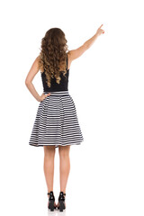 Woman In Striped Skirt And High Heels Is Pointing Up, Rear View.