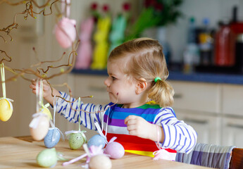 Cute little toddler girl decorating tree bough with colored pastel plastic eggs. Happy baby child having fun with Easter decorations. Adorable healthy smiling kid in enjoying family holiday