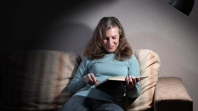 Woman reading a book while sitting at home on the sofa under the light of the lamp