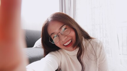 Beautiful Asian woman wearing glasses is smiling and waving to the video camera for a selfie to a...