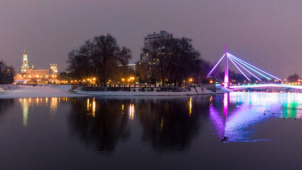 Bridge (Mist Zakokhanykh) across river in illuminated Skver Strilka in Kharkiv city center. Winter aerial evening colorful photo with water reflection
