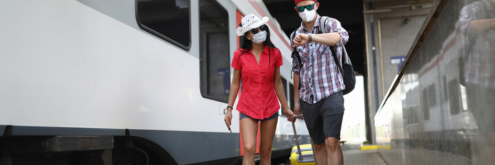 A man and a woman go with a suitcase to board the train. Medical masked travelers walk the train station