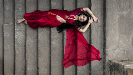 Portrait of beautiful Indian girl in heritage stepwell wearing traditional Indian red saree, gold jewellery and bangles. Maa Durga agomoni shoot concept. Traditional woman on stairs with poses