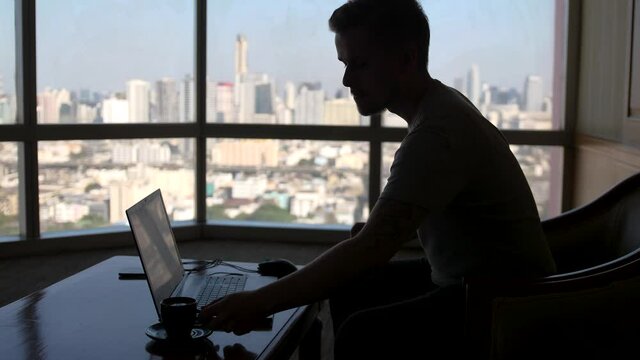 A concentrated young businessman is working on a laptop computer using his notebook sitting by the window with urban view and drinks coffee
