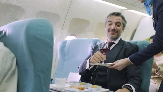 Senior businessman or passenger listen music with earphone, sitting in the first class of airplane. Enjoying the flight and drinking red wine, feeling smile and happy. Stewardess serve food and drink