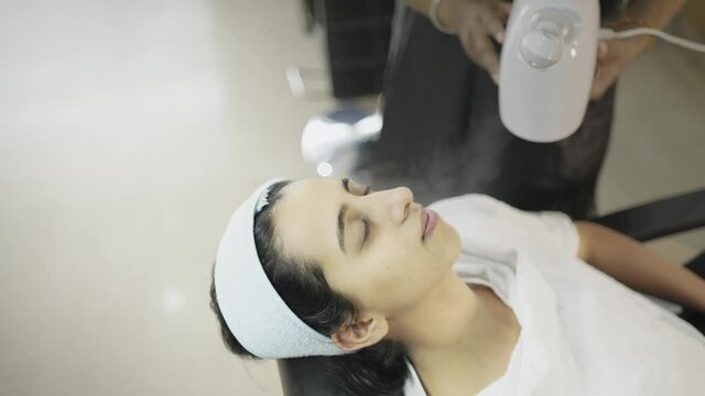 close view shot of a young indian asian female lying on a beauty salon recline chair in spa with eyes closed where the staff person is giving warm steam thoroughly on her face with facial steamer