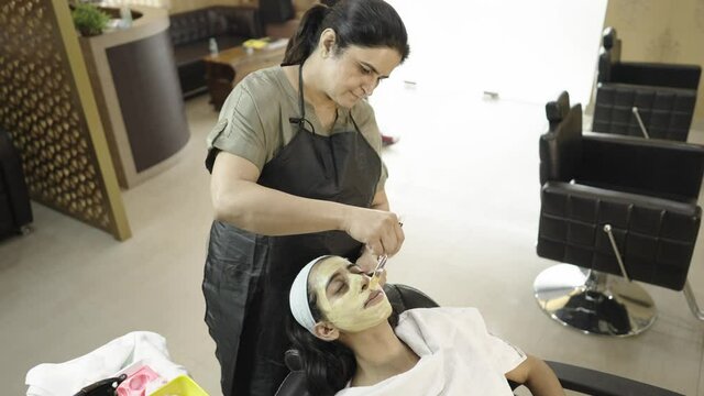 far view shot of a beautician applying face mask cream using a brush on the face of a young indian asian female sitting in a comfy chair in a beauty parlour