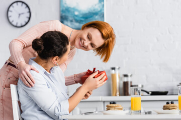 cheerful lesbian woman presenting gift on valentines day to african american lesbian girlfriend in kitchen