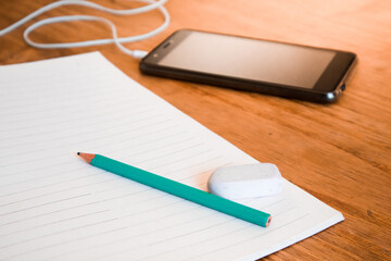 Pencil, eraser, notebook, cell phone and headphones on wooden table in daylight